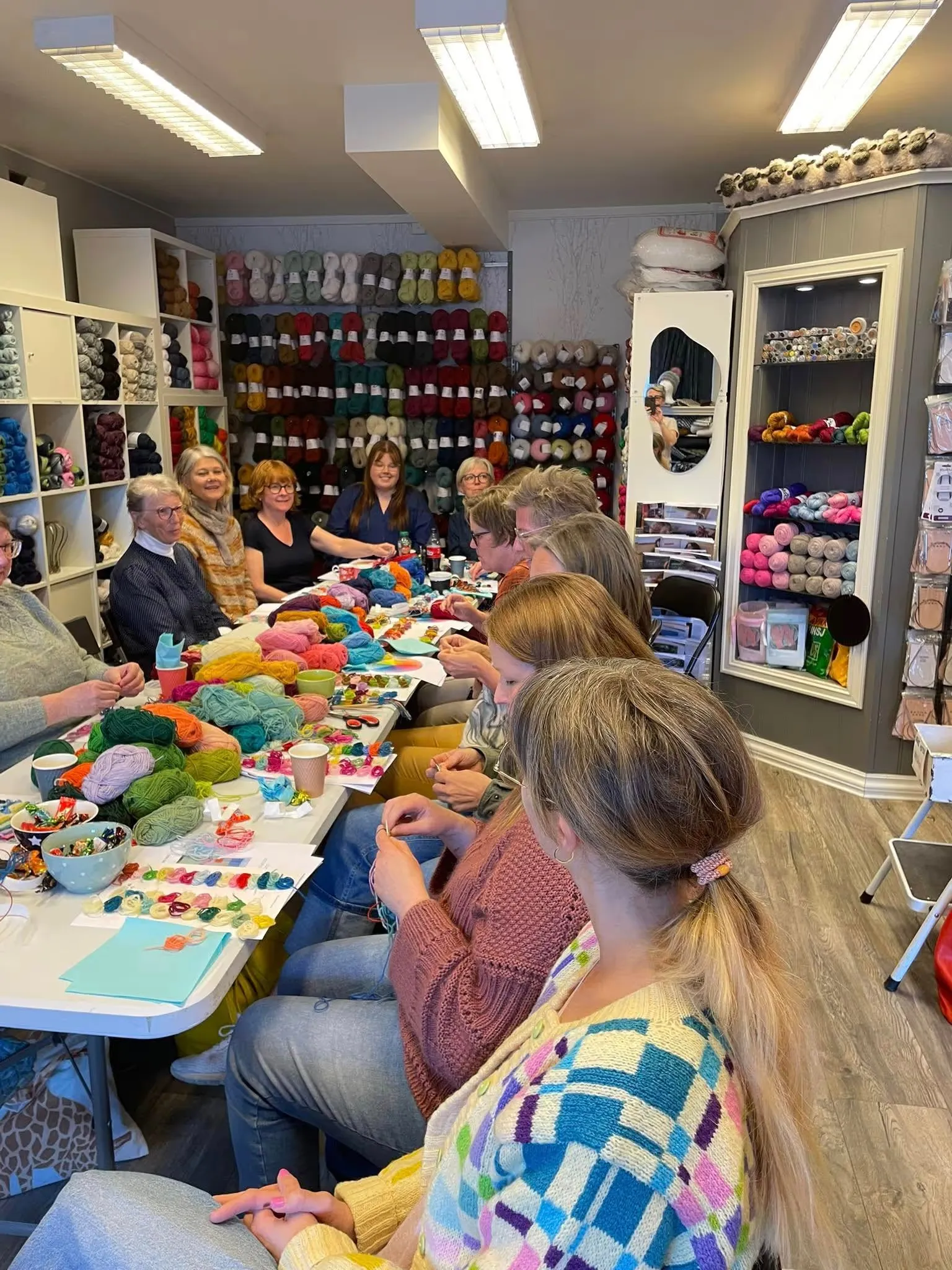 Strikkekafé rundt et langbord i butikken, med garn i hyllene bak
